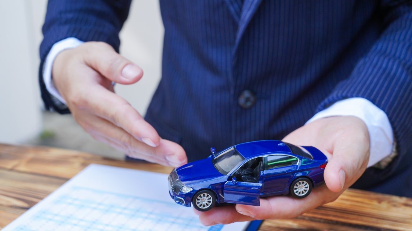 A man in a blue suit showing off a toy blue car over a spreadsheet.